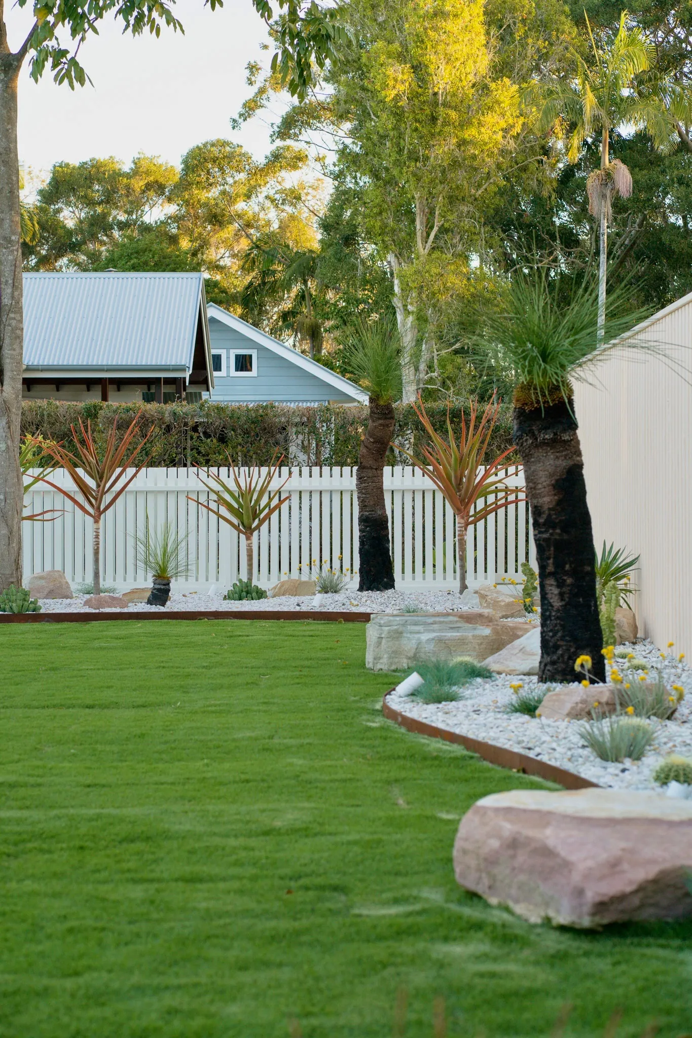 New turf and a sandstone boulder beside a pebble bed under a palm, edged in corten